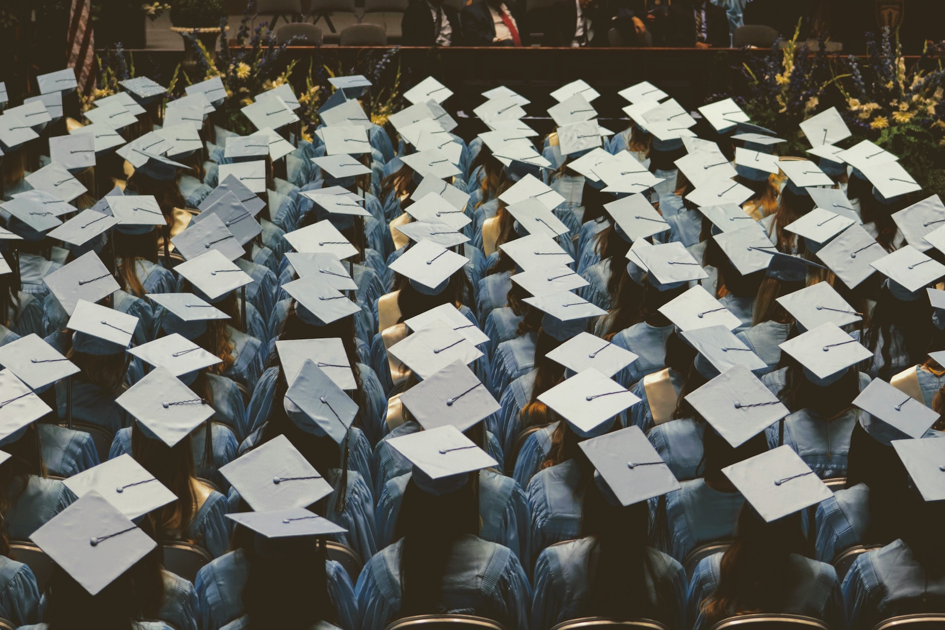 A multitude of rows of graduates in their caps and gowns sit in an auditorium, facing the stage.