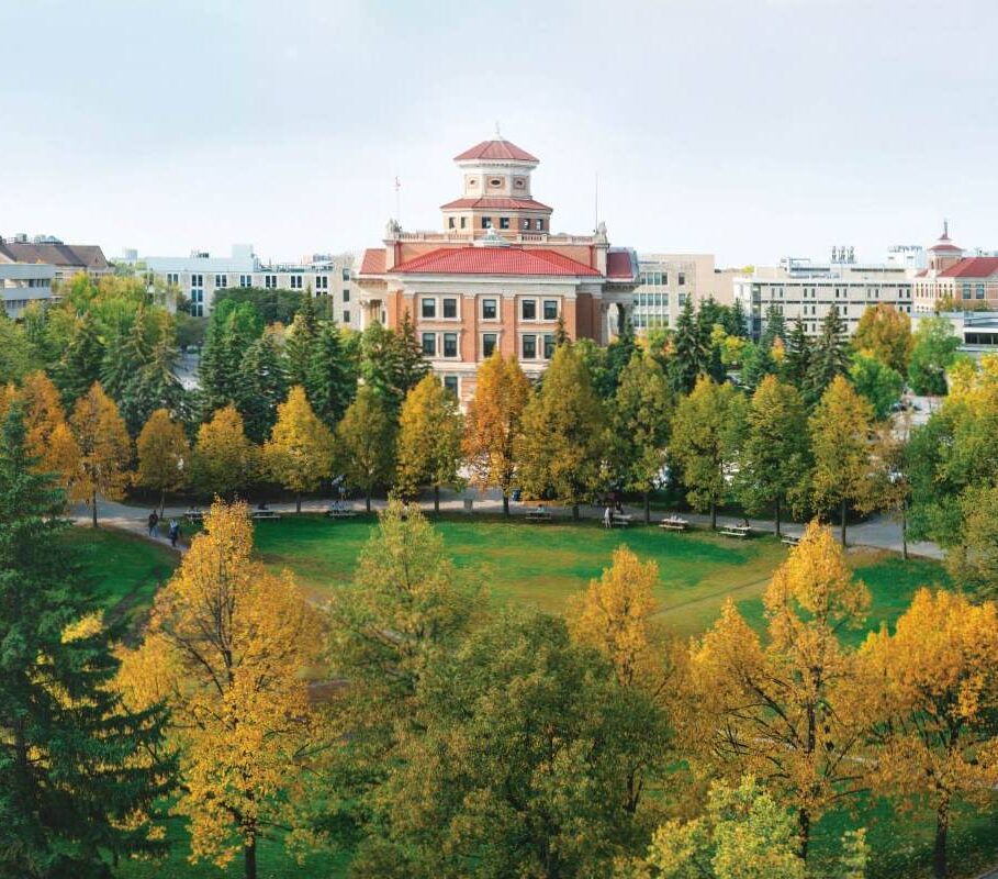 Un imposant bâtiment administratif en briques rouges se trouve derrière une cour entourée d'arbres qui changent de couleur à l'automne.